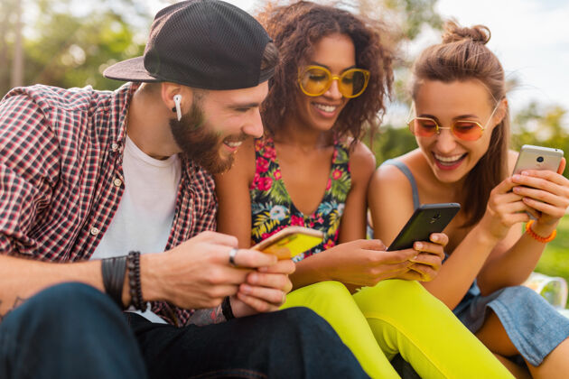 时尚快乐的年轻朋友们坐在公园里用智能手机微笑 男人和女人一起玩人无线情绪
