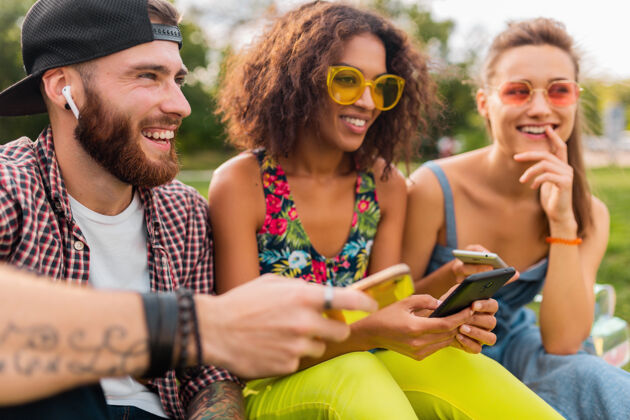 团体快乐的年轻朋友们坐在公园里用智能手机微笑 男人和女人一起玩年轻耳机情绪