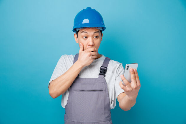 电话关心的年轻建筑工人戴着安全帽 穿着制服 手捂着嘴看着手机保持制服安全