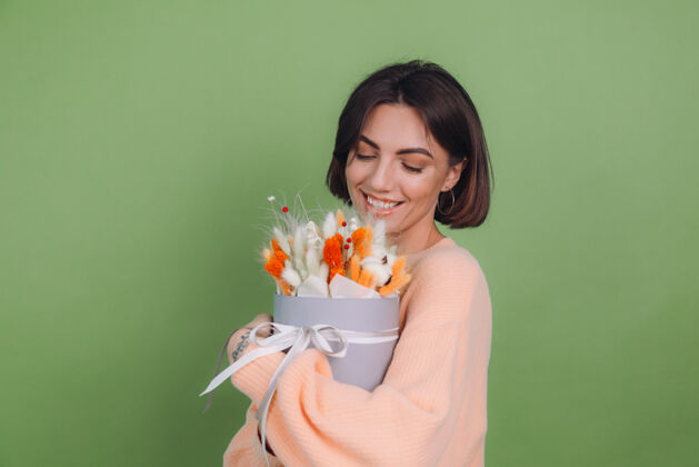 花年轻女子穿着休闲桃色毛衣隔离在绿橄榄墙上手持橙白色花盒组成的棉花 吉普赛拉小麦和拉古鲁斯作为礼物开心惊喜干花女性魅力