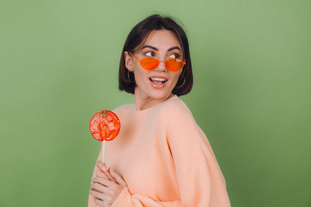化妆品年轻时尚的女士穿着休闲的桃色毛衣 戴着橙色的眼镜 隔离在绿色橄榄色的墙上 带着橙色棒棒糖的正面微笑复制空间休闲食物女孩