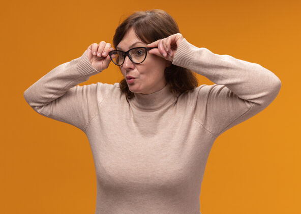 高领毛衣穿着米色高领套头衫 戴着眼镜的中年妇女站在橘色的墙上近距离地看向一边穿封闭站