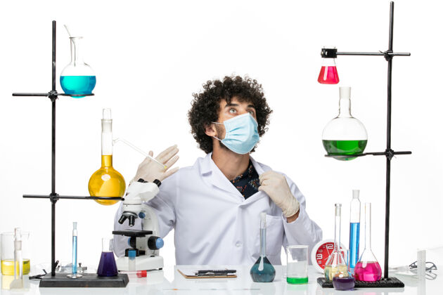 面具前视图男性化学家穿着医疗服 戴着面罩 坐在浅白的空间里 拿着溶液男性化学家实验室烧杯