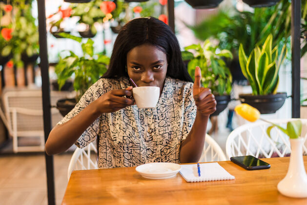 时尚成功的女人竖起大拇指非洲女人拿着咖啡杯 在咖啡馆微笑着竖起大拇指手掌等待非洲