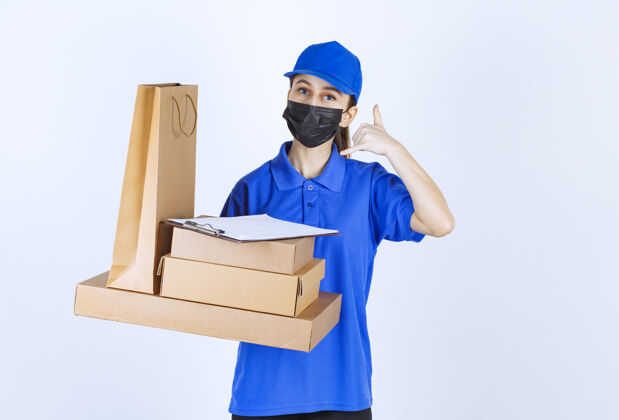 食品带着面具和蓝色制服的女快递员拿着一个纸板购物袋和多个箱子 一边要求打电话员工预订送货