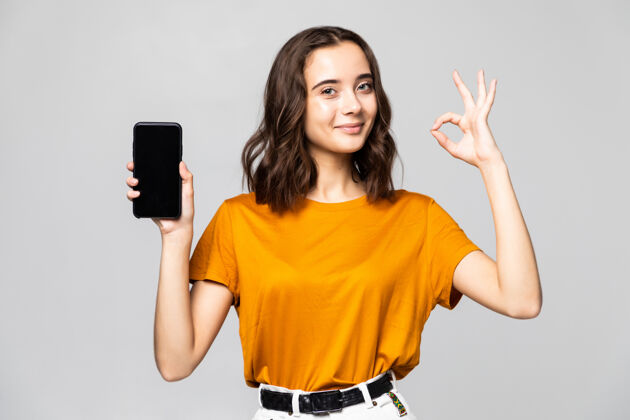 持有穿着休闲服的快乐女人在灰色的墙壁上展示了一个空白的智能手机屏幕和一个好的手势脸女士打开