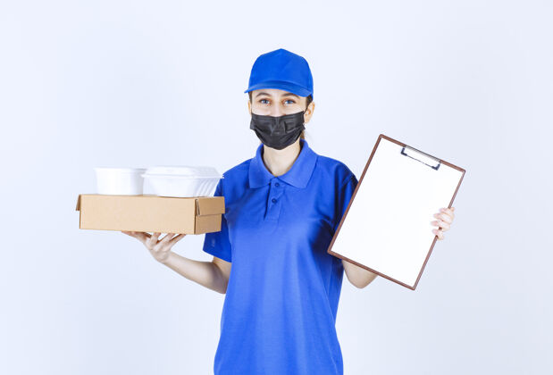 年轻人女信使戴着口罩 身穿蓝色制服 手里拿着一个纸板箱 外卖包裹 并出示清单供签字促销聪明人员工