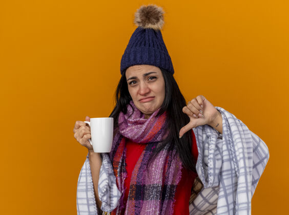 疾病年轻的白种病女孩戴着冬天的帽子 围巾裹着格子布 手里拿着一杯茶 拇指朝下 被隔离在橙色的墙上 留着复印空间茶橙色杯子