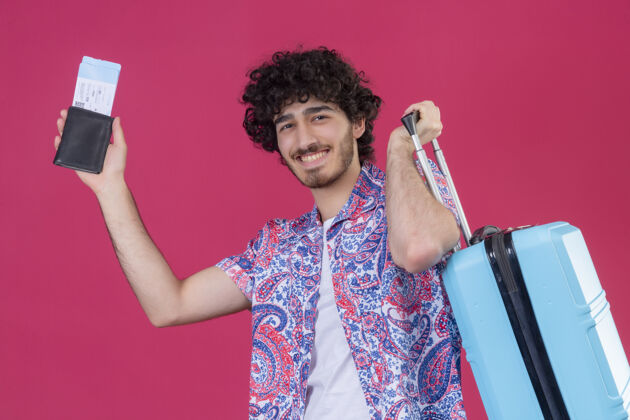 男人微笑着的年轻英俊的卷发旅行者 手里拿着钱包 机票和手提箱 孤零零地站在粉红色的墙上钱包卷发帅哥