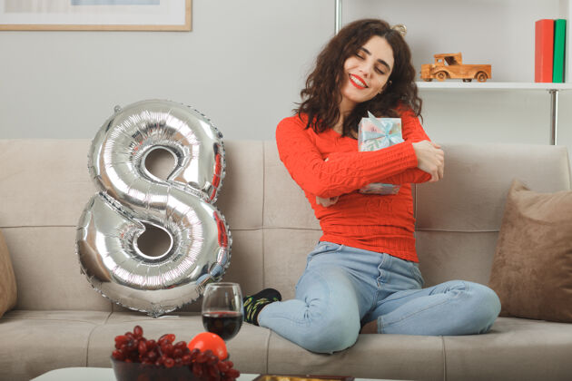 房间快乐快乐的年轻女子穿着休闲服 兴高采烈地微笑着坐在沙发上 拿着8号形状的气球拥抱礼物庆祝3月8日国际妇女节沙发气球坐着