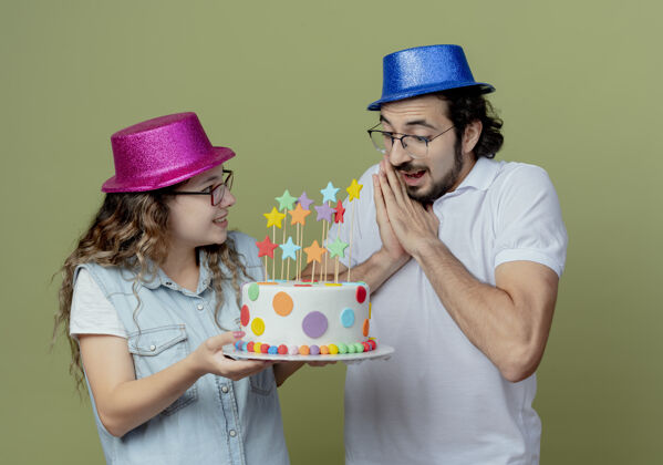 男人一对戴着粉蓝色帽子的年轻夫妇高兴地把生日蛋糕送给了被隔离在橄榄绿墙上的惊讶的家伙给帽子年轻