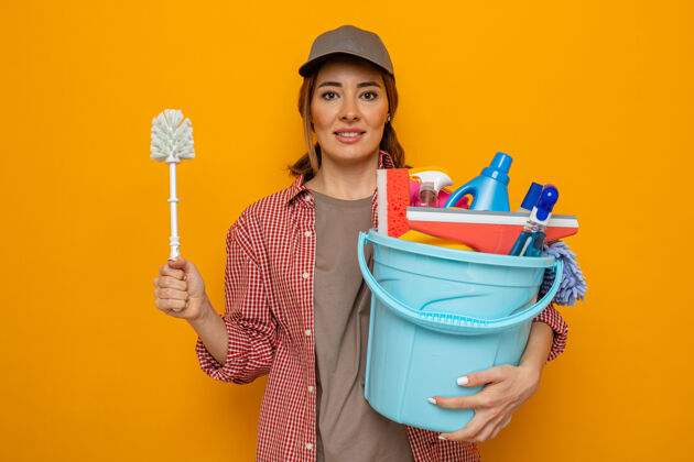 工具身穿格子衬衫 头戴鸭舌帽的年轻清洁女工 手里拿着清洁工具和清洁刷 面带微笑 快乐而积极地准备好了抱着格子年轻