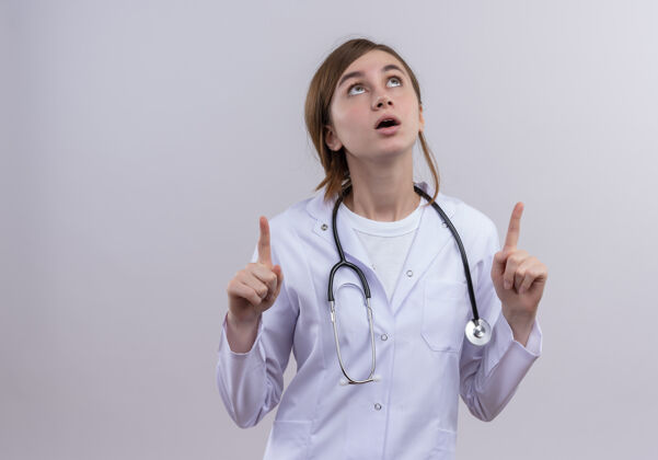 听诊器穿着医用长袍和听诊器的年轻女医生惊讶地抬起头来 在有复印空间的白色隔离墙上竖起手指手指年轻人升起