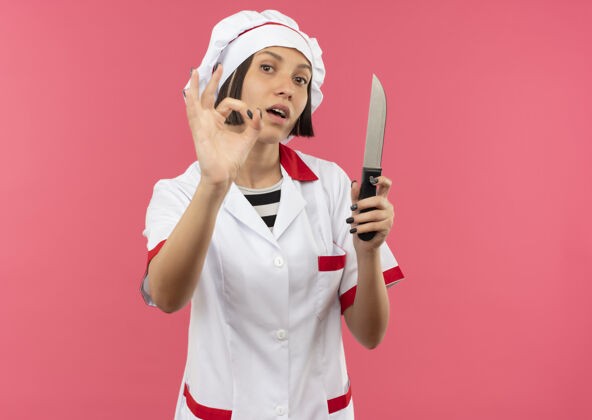 标志自信的年轻女厨师穿着厨师制服拿着小刀 做着ok的标志 隔离在粉色的复制空间里女持有烹饪