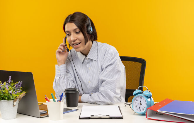 年轻印象深刻的年轻呼叫中心的女孩戴着耳机坐在办公桌上把手放在耳机上孤立的橙色坐着电话印象