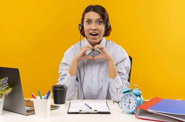 办公桌印象深刻的年轻呼叫中心的女孩戴着耳机坐在办公桌上做心脏孤立的橙色标志标志印象电话