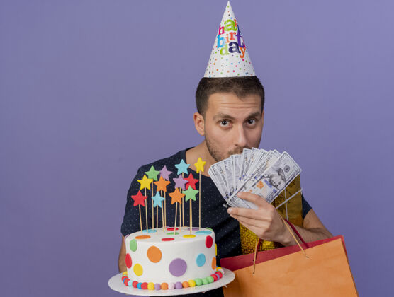 惊喜戴着生日帽的白人帅哥拿着birhtday蛋糕纸袋礼品盒和钞票 紫色背景上有复制空间蛋糕礼物钱