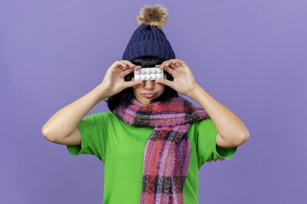 持有年轻生病的白人女孩戴着冬天的帽子和围巾 手里拿着一包药片 眼前一片紫色的背景孤立包药片女孩