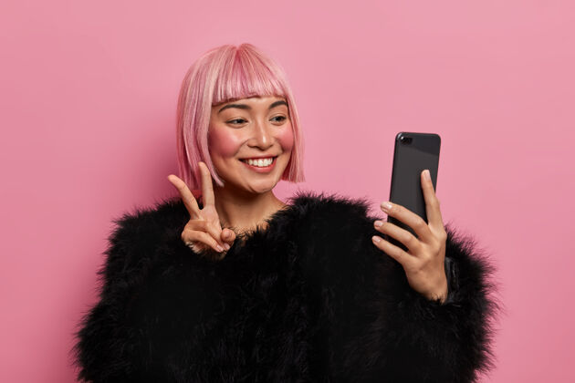 正面人 科技 生活方式的概念微笑快乐的粉红色头发的女人穿着蓬松的黑色毛衣 自拍 显示v标志或和平的姿态 发送良好的积极氛围 享受在线视频通话牙关示范视频