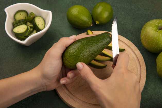 小吃俯视图：女性用刀在木质厨房板上切一个鲜绿色的鳄梨 绿色的墙壁上隔离着酸橙和绿色的苹果顶部板配料
