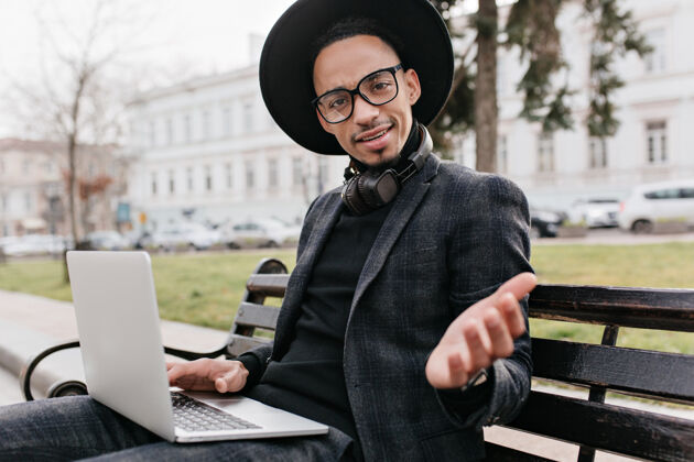 工人非洲自由职业者在广场上用笔记本电脑工作时表示惊讶一张惊讶的黑人戴着帽子 坐在长凳上拿着电脑的户外照片西装休闲男人