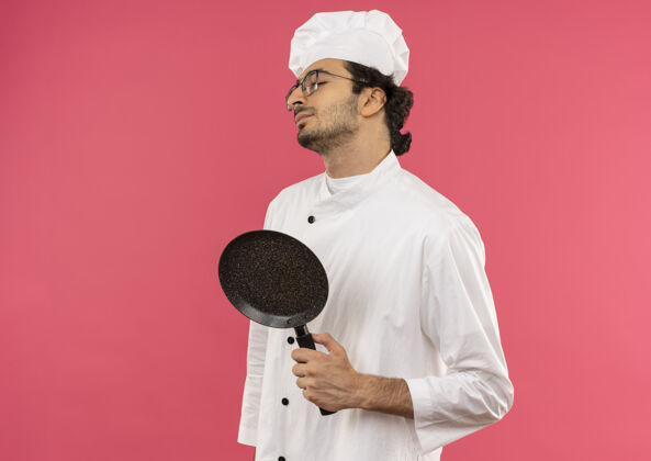制服年轻的男厨师闭着眼睛 穿着厨师制服 戴着眼镜 手里拿着粉红色的煎锅封闭薯条烹饪