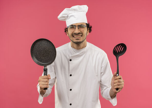 粉色压力很大的年轻男厨师穿着厨师制服 戴着眼镜 手里拿着平底锅和粉红色的抹刀年轻制服平底锅
