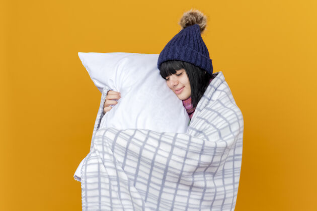 女孩年轻生病的高加索女孩戴着冬天的帽子和围巾 裹着格子呢 抱着枕头 用它抚摸着脸 闭着眼睛 在橙色的背景上 留着复制空间年轻穿帽子