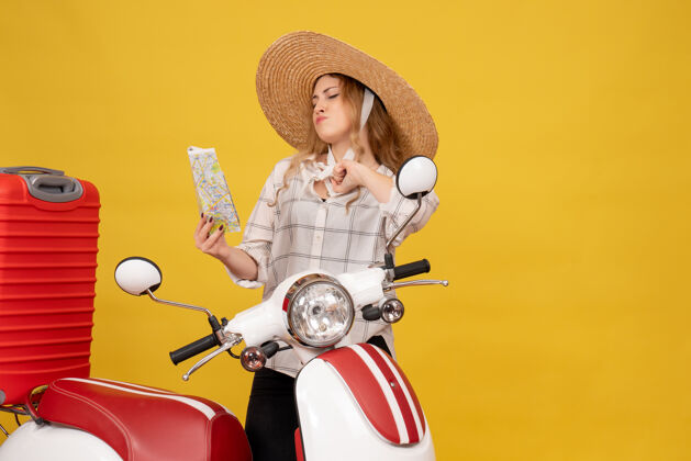 坐着头戴帽子的骄傲的年轻女子坐在摩托车上拿着地图拿着她的行李车辆地图摩托车