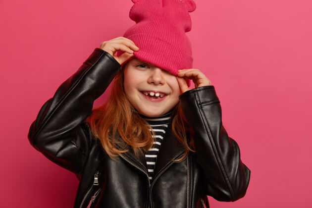 帽子剪短的小女孩形象从帽子上看 掩面 穿着时髦的黑色皮夹克 穿着时髦的衣服有积极的雄心壮志的样子孤立在粉红色的墙上孩子 情感 风格狐狸女孩雄心勃勃