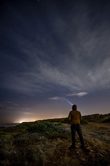 颜色一个人站在岩石上 看着夜晚闪亮的星星的垂直镜头夜人站立