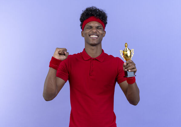 手势年轻的美国黑人运动男子戴着头带和腕带 紧闭双眼 拿着优胜者杯 在蓝色的舞台上表现出“是”的姿态蓝色杯子冠军