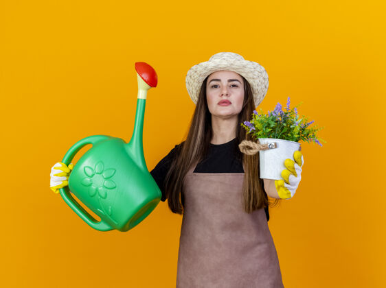花自信美丽的园丁女孩穿着制服 戴着园艺帽 戴着手套 举着浇灌罐 拿着花盆里的花 对着隔离在橙色背景下的摄像机信心制服帽子