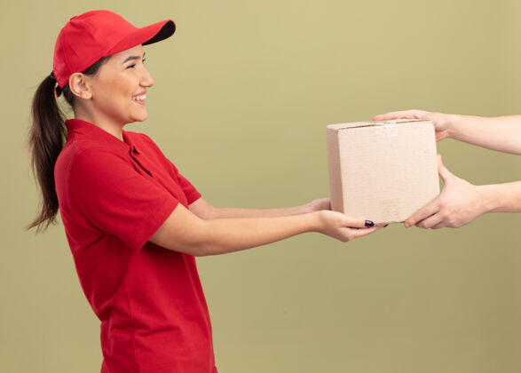 制服身着红色制服 头戴鸭舌帽的年轻女送货员站在绿色墙壁上 微笑着向顾客赠送纸板箱女人站立年轻人