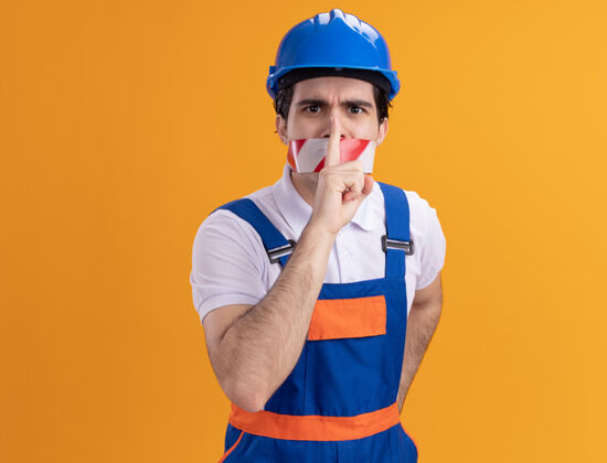 胶带年轻的建筑工人穿着建筑制服 戴着安全帽 嘴上缠着胶带 站在橘色的墙上 用手指捂着嘴唇 注视着前方 做着沉默的手势安全男人站立