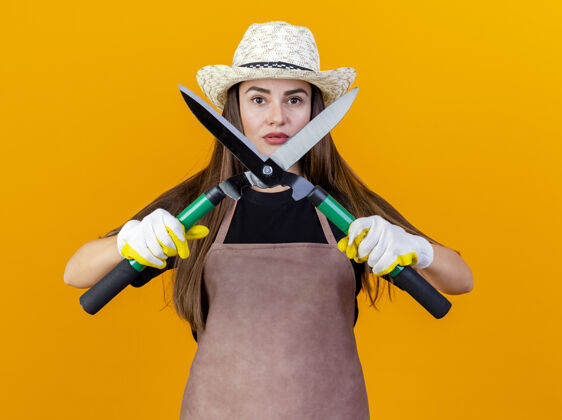 自信自信美丽的园丁女孩穿着制服 戴着园艺帽 戴着手套 拿着剪子 在橙色背景上隔离开来女孩花园制服