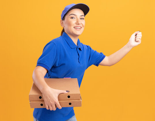 制服身着蓝色制服 头戴鸭舌帽的快乐的年轻送货员正在橘色的墙上为顾客运送比萨饼盒顾客跑步年轻人