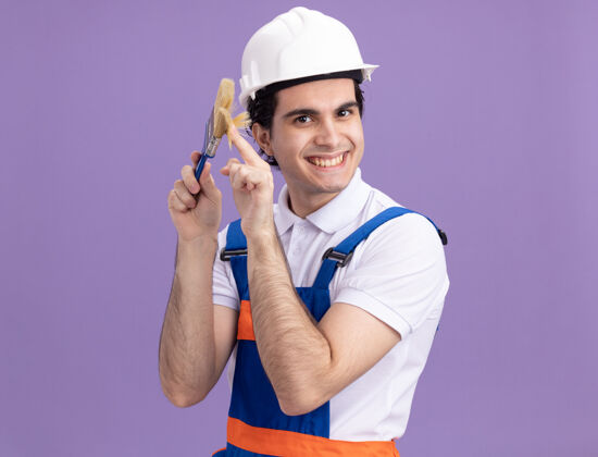 头盔快乐的年轻建筑工人身穿建筑制服 戴着安全帽 拿着油漆刷 站在紫色的墙上 面带微笑地看着前方抱男人站