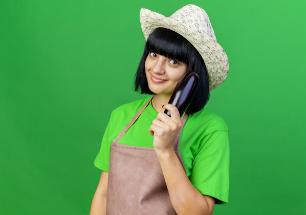 持有高兴的年轻女园丁穿着制服戴着园艺帽拿着茄子花园茄子绿色