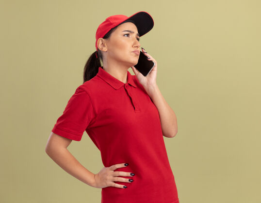 手机身穿红色制服 戴着帽子的年轻女送货员站在绿色的墙上讲着手机 看上去很困惑送货电话女人