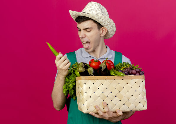 男性年轻的男园丁戴着园艺帽 伸出舌头 拿着菜篮子和辣椒穿辣椒粉红色