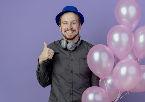 帽子微笑的帅哥 戴着蓝色帽子 脖子上戴着耳机 站着氦气球 竖起大拇指孤立地站在紫色的墙上男人耳机向上