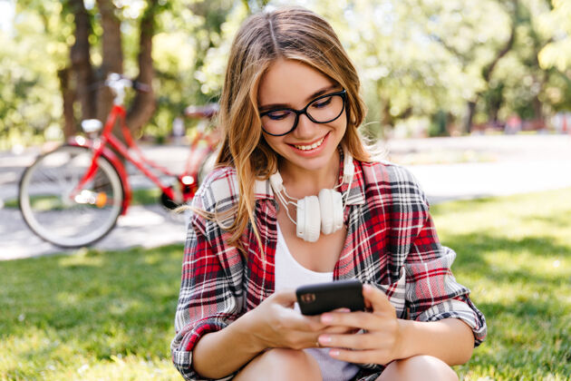 女人浪漫的女孩在美丽的公园里休息时发短信一张快乐的金发女人拿着智能手机坐在草地上的户外照片树可爱自然