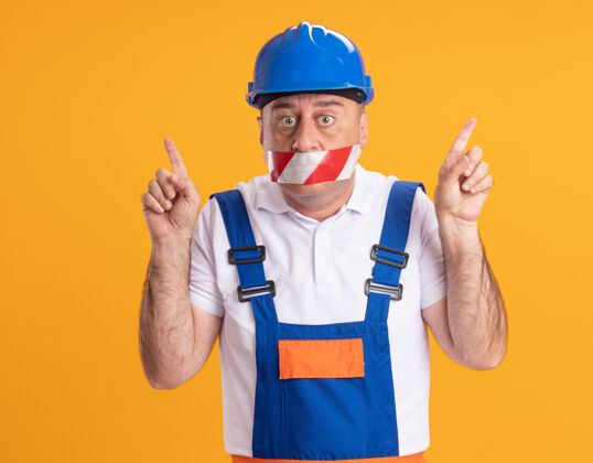 胶带震惊的白人成年建筑工人穿着制服 用胶带捂住嘴巴 两手指着橙色人建筑工人掩护