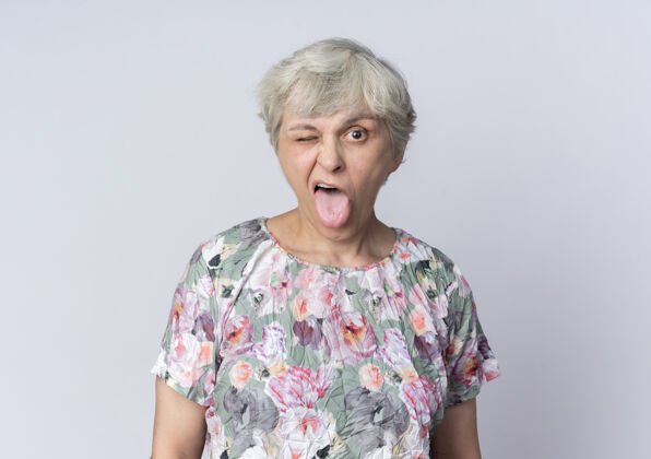 眼睛快乐的老妇人眨着眼睛 伸出舌头孤立地站在白墙上老人女人舌头
