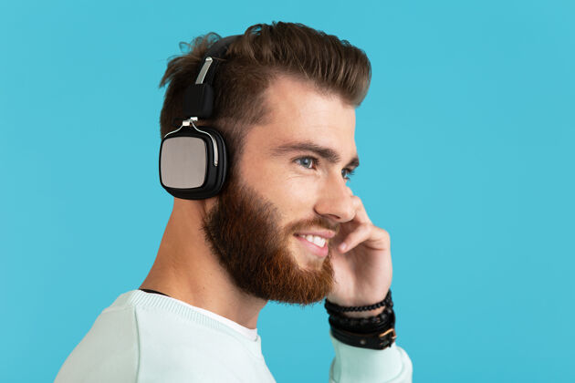 音频时尚迷人的年轻胡须男子的肖像在无线耳机上听音乐现代风格自信的情绪在蓝色背景上孤立城市家伙享受