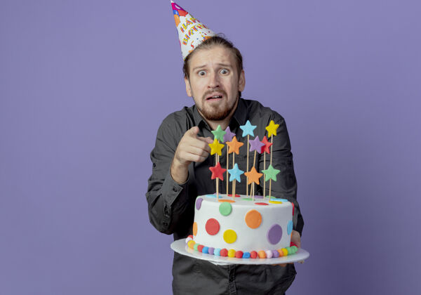 蛋糕戴着生日帽的焦急帅哥拿着蛋糕 孤立地指着紫色的墙上向前点着急