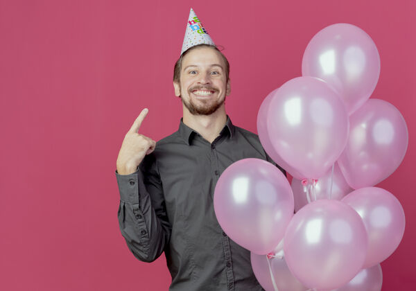 氦戴着生日帽的快乐帅哥站在那里 氦气球指着粉色墙上孤立的帽子气球快乐站着