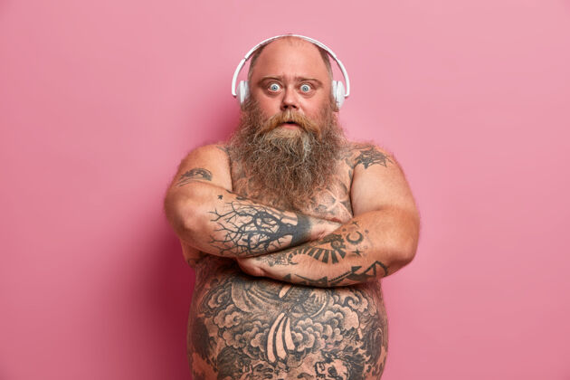 腹部奇怪的胡须男人的肖像保持双臂合拢 用的眼睛看 买了耳机在假日销售 捕捉积极的氛围 有裸体纹身肚子 肥胖 因为懒惰的生活方式男人情绪音乐