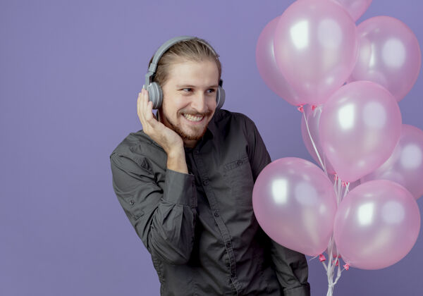 紫色高兴的帅哥戴着耳机拿着氦气球看着隔离在紫色墙上的一面气球帅哥氦气